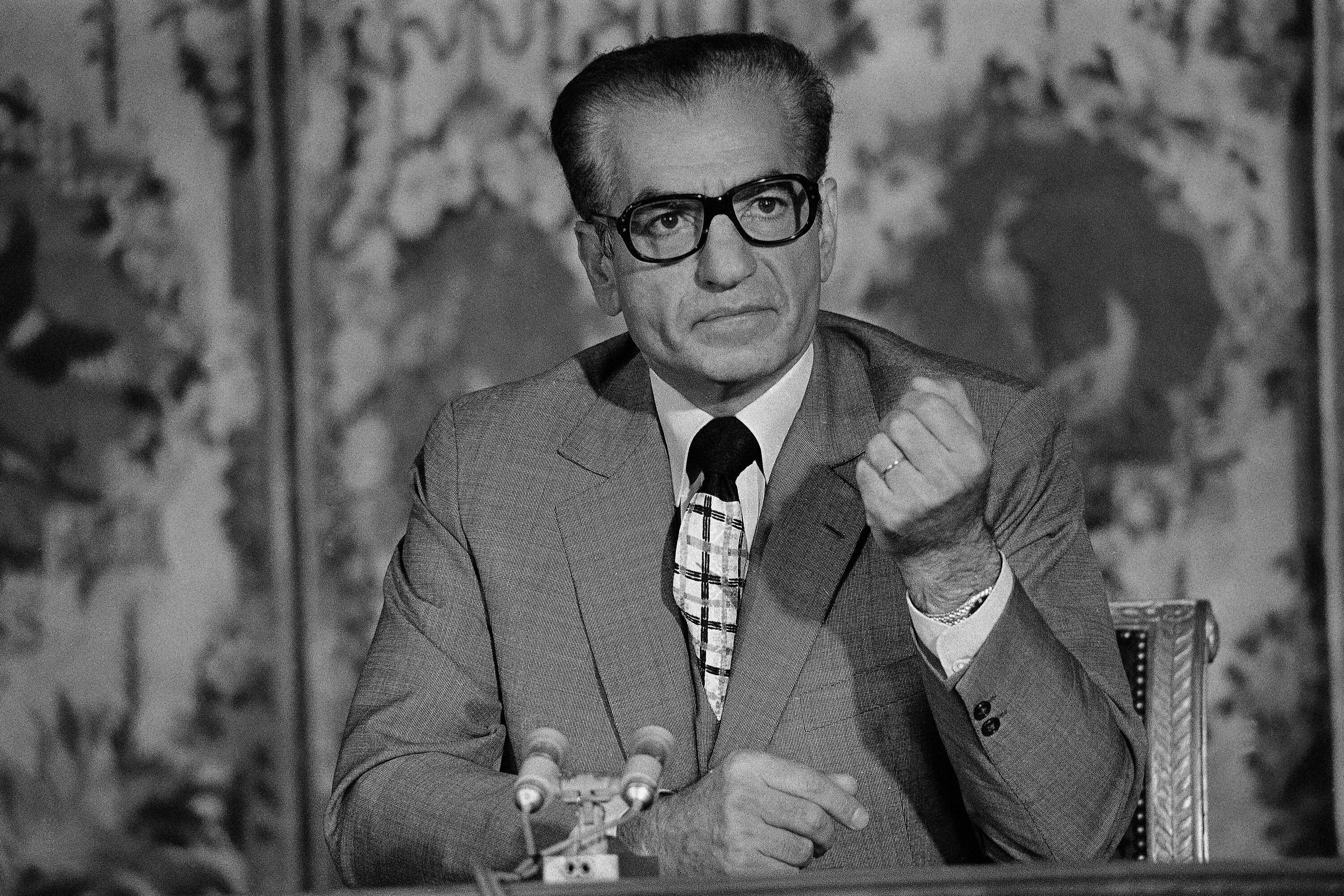 Man in suit wearing thick rimmed glasses holds up fist.
