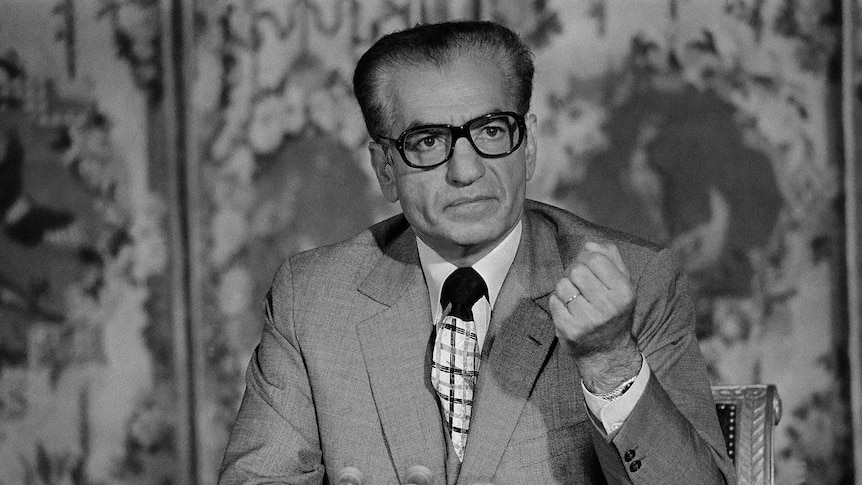 Man in suit wearing thick rimmed glasses holds up fist.