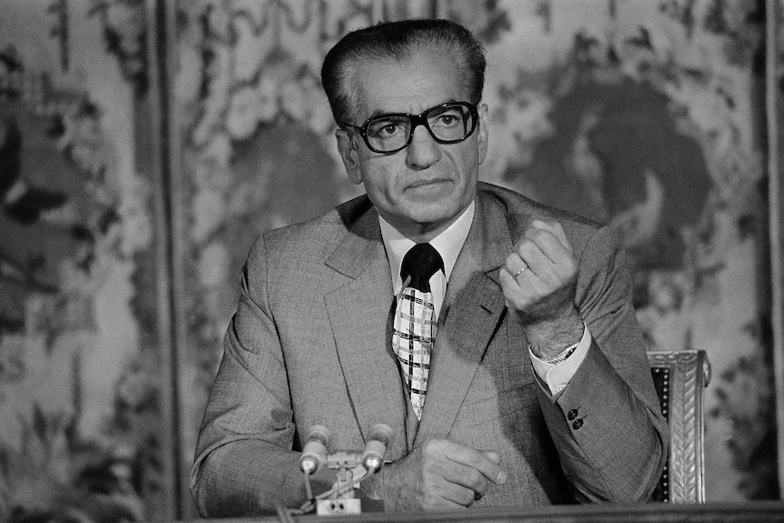 Man in suit wearing thick rimmed glasses holds up fist.