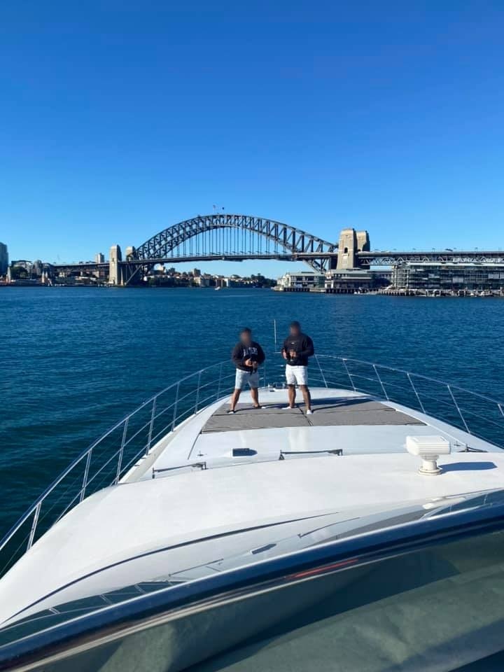 Two men stand on the bow of a large boat with Sydney Harbour Bridge in the background.