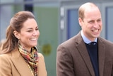 Kate and Prince William smile, not looking directly at the camera. They're both wearing brown coats. 