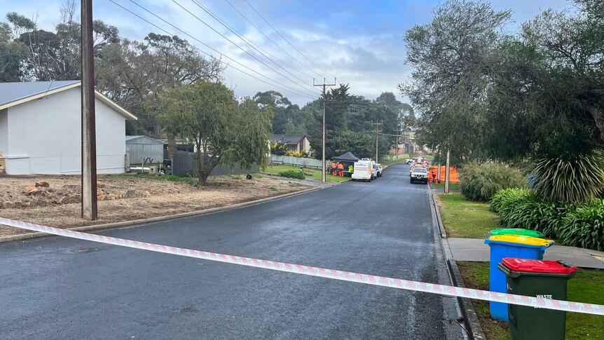 A suburban street with orange and white tape blocking off an area, with emergency services vehicles outside a house