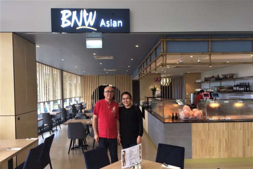 A man and a woman stand among the tables in a restaurant under a sign that says BNW Asian.