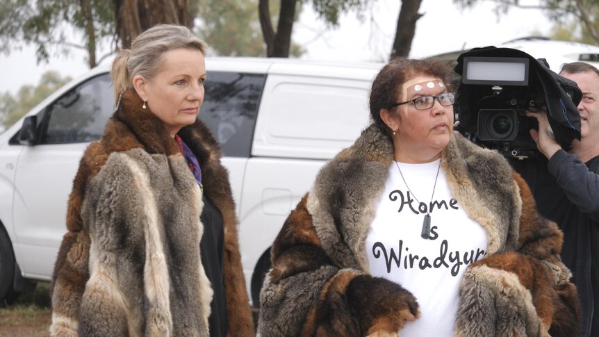 Two women wear possum skin cloaks in front of a white van and a man holding a TV camera.