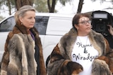 Two women wear possum skin cloaks in front of a white van and a man holding a TV camera.