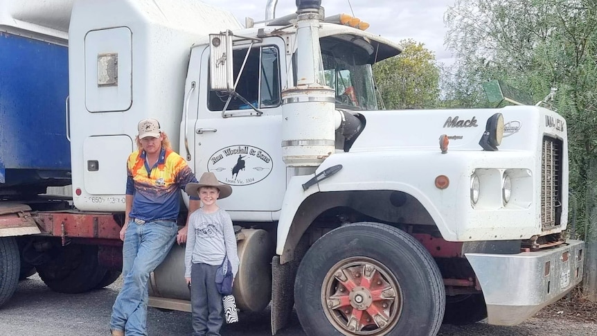 A man in jeans, boots and a work shirt leans against a large truck alongside a smiling young boy wearing an Akubra.
