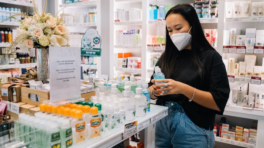 A young woman wearing a face mask stands in a pharmacy holding a bottle of hand sanitizer.