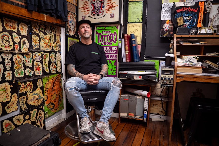 A man sits in the corner of a studio, with tattoo memorabilia on the walls around him.