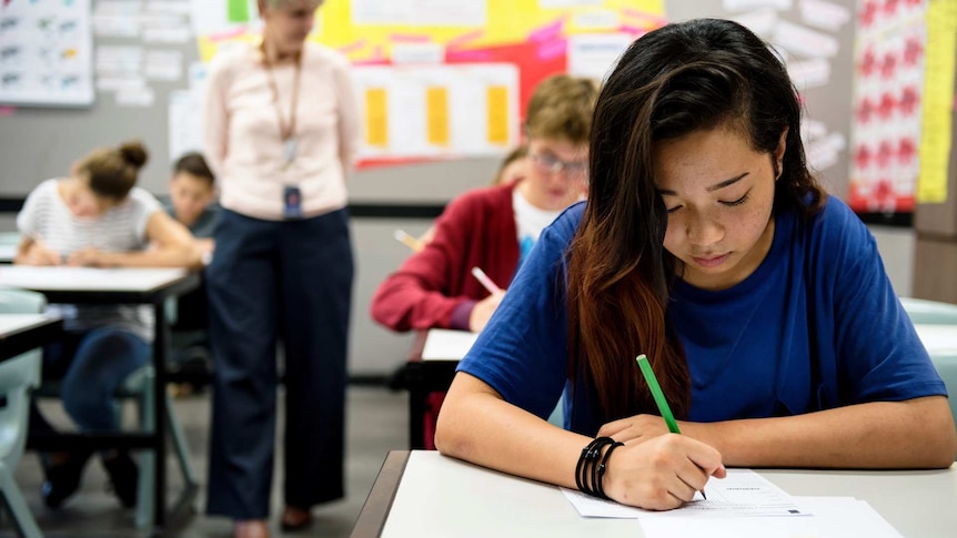 An Asian high school girl writing on paper at a school desk. There are other students at desks around her doing the same.