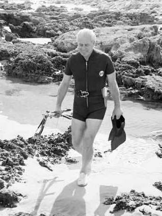 A man in a short wetsuit walks to the water carrying flippers and fishing spears.