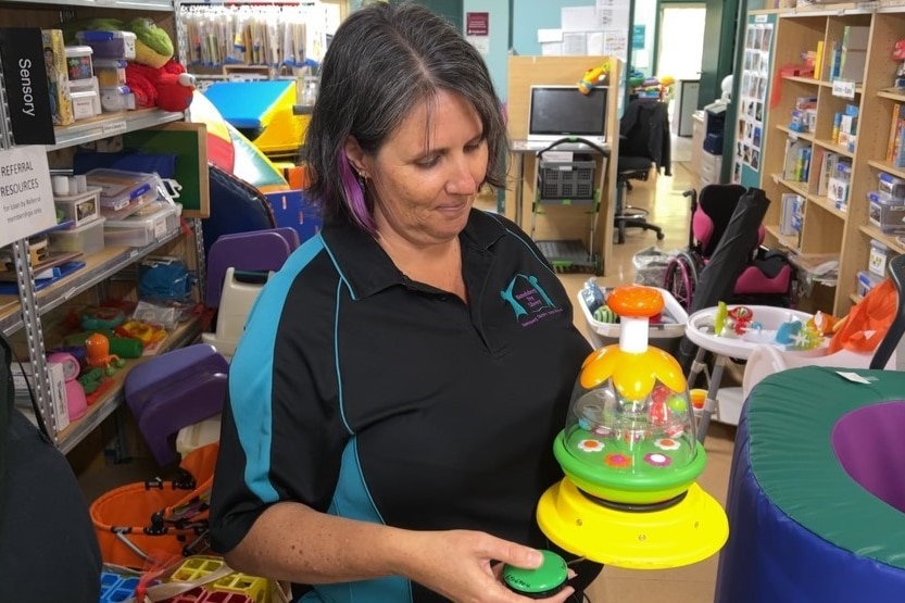 A woman in a polo shirt and grey and purple hair looks down at a colourful toy