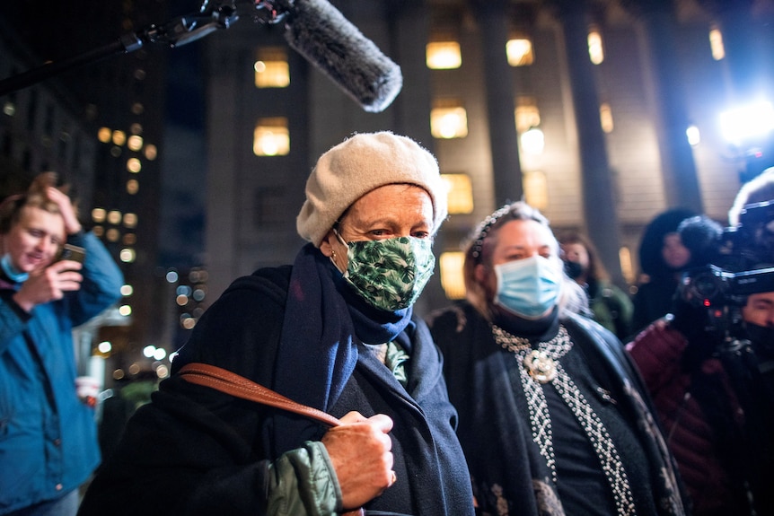 An older woman wearing a cream beret, navy cardigan and patterned face mask is surrounded by photographers