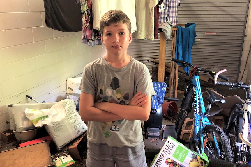 Year 8 student Jeff Dawson stands, arms crossed in a garage surrounded by water-damaged belongings.