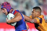 A Newcastle Knights NRL players carries the ball with his left arm as he is tackled from behind by Brisbane Broncos opponents.