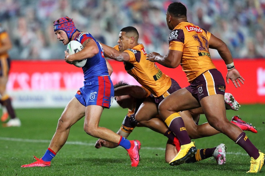 A Newcastle Knights NRL players carries the ball with his left arm as he is tackled from behind by Brisbane Broncos opponents.