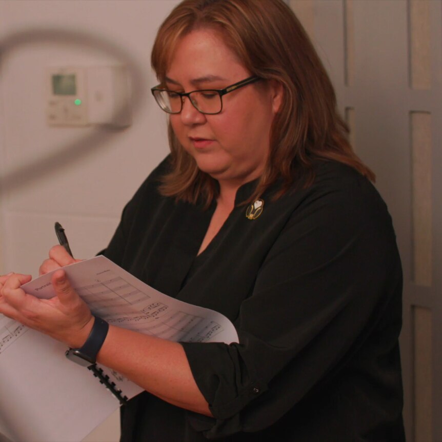 Composer Jessica Wells stands next to a wall looking down at a music score. An out of focus microphone is in the foreground.