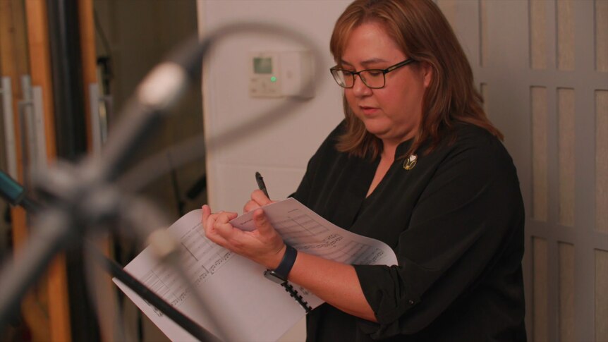 Composer Jessica Wells stands next to a wall looking down at a music score. An out of focus microphone is in the foreground.