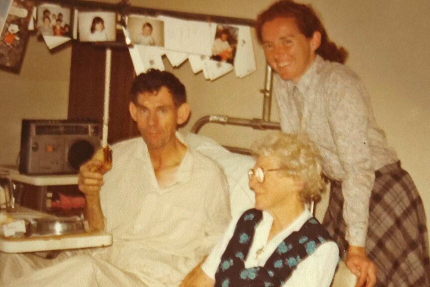 A man eating a sandwich in a hospital bed with relatives around him.