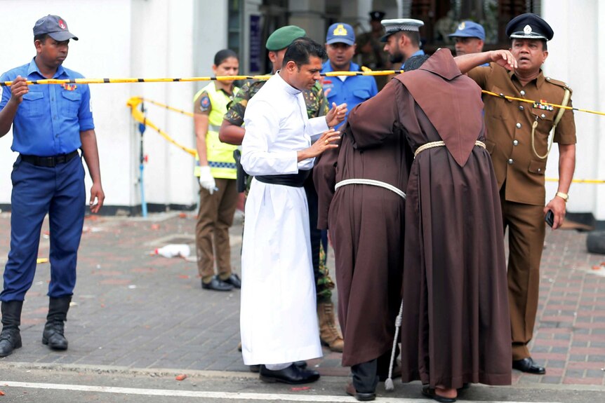 Two men in brown religious robes walking under a yellow tape towards a group of men in police uniforms.