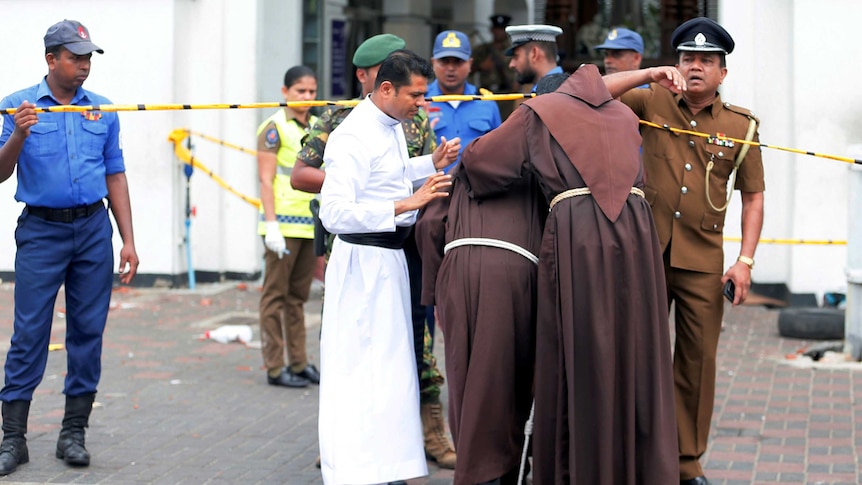 Two men in brown religious robes walking under a yellow tape towards a group of men in police uniforms.