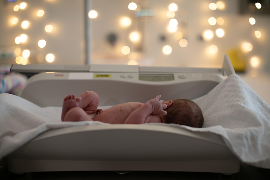 A newborn baby lies scrunched up on a changetable, looking toward a wall decorated with fairy lights.