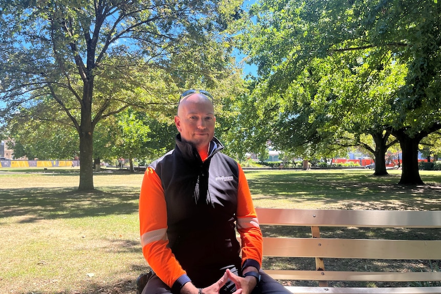 A bald man with sunglasses on his head wearing an orange high-vis top and black vest sits in a park.
