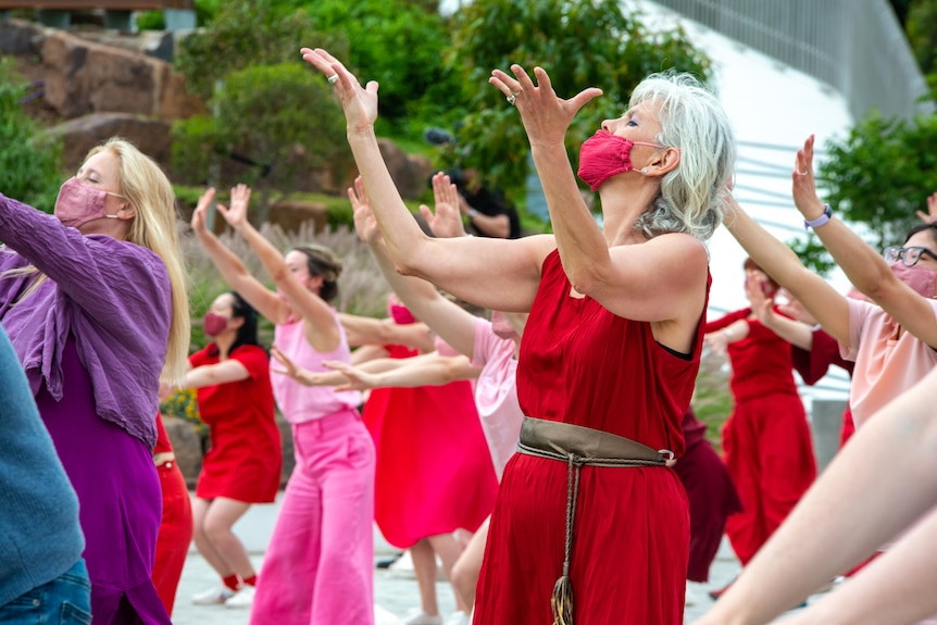 A large group of dancers dressed in block colours and wearing masks stretch out their arms in synchronised movement outdoors.