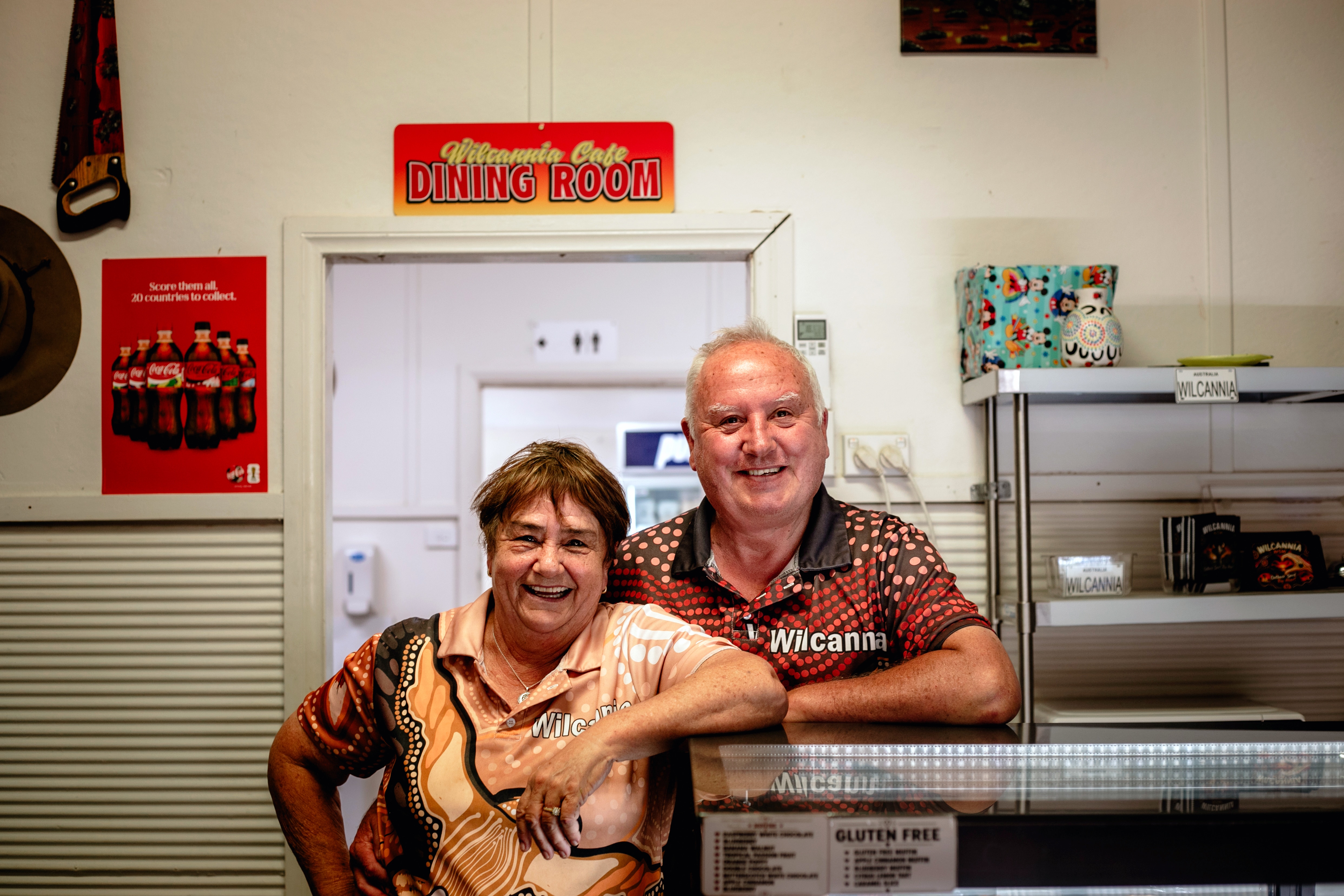 Shona and Conrad Cook, an older couple, laugh as they stand by a bench in a cafe.o