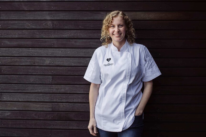 Amber Kaba wearing white chef jacket and standing in front of wall made of wood.