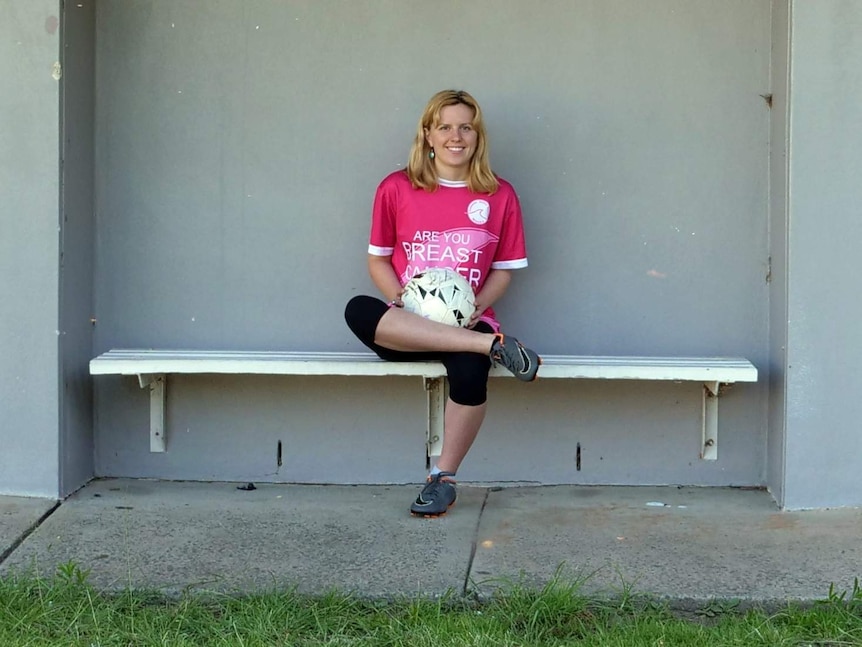 A woman sits with a soccer ball on her lap in front of a wall