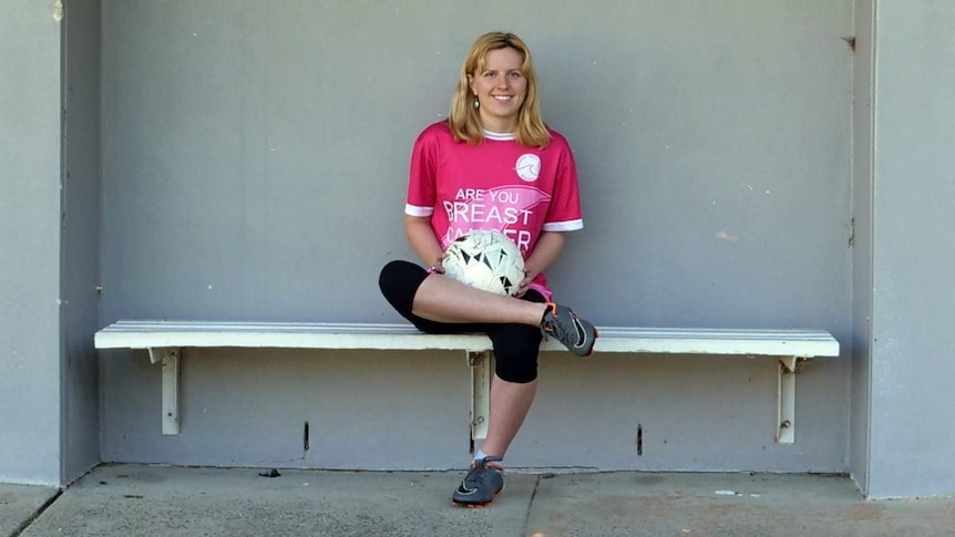A woman sits with a soccer ball on her lap in front of a wall