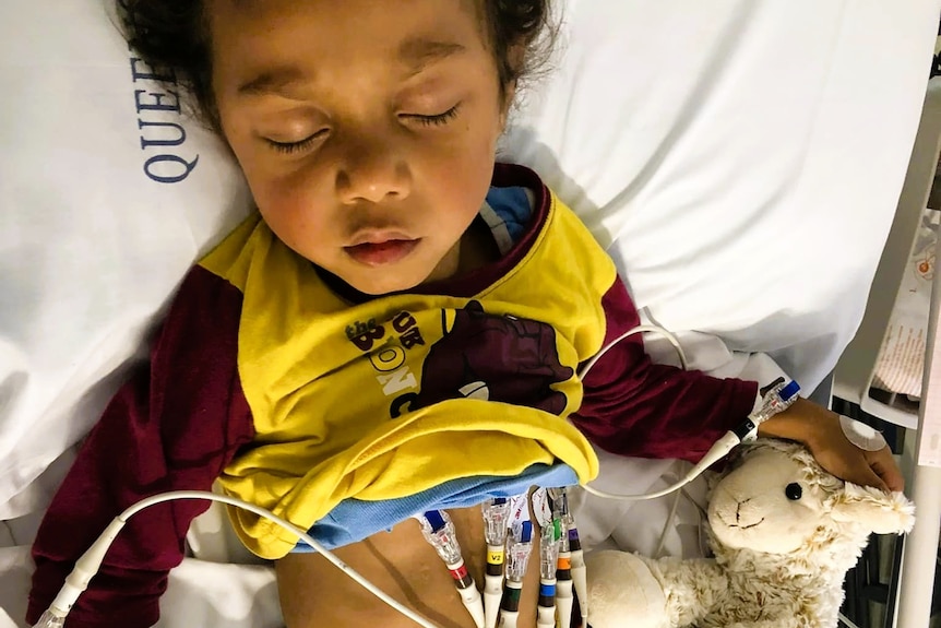 A young boy sleeping in a hospital bed, wearing Broncos PJs and with numerous wires and chords on his body.