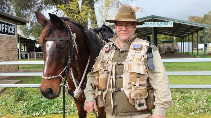A man dressed in army regalia stands next to a horse in a paddock.