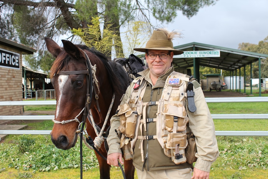 A man dressed in army regalia stands next to a horse in a paddock.