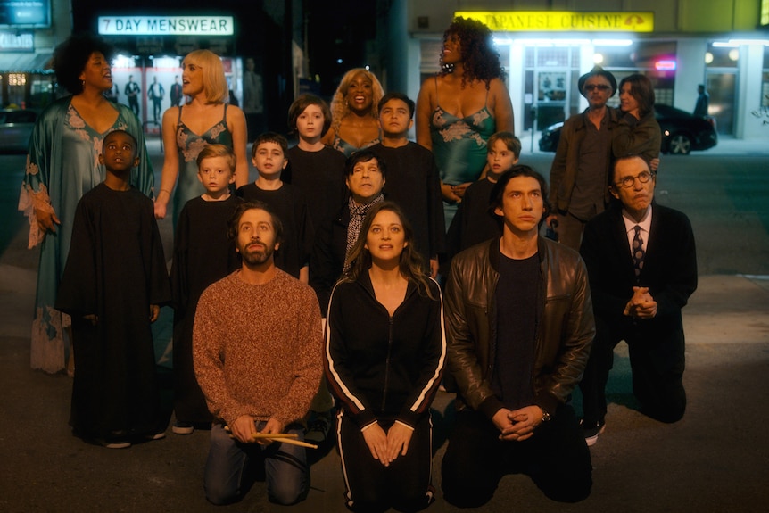 Simon Helberg, Marion Cotillard and Adam Driver kneel in front of an animated group of singers under a streetlamp, looking up.