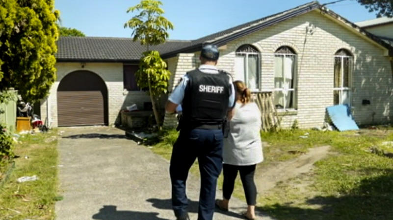 Man in blue and black uniform with Sheriff written on back walks next to a woman down a driveway toward a cream brick house