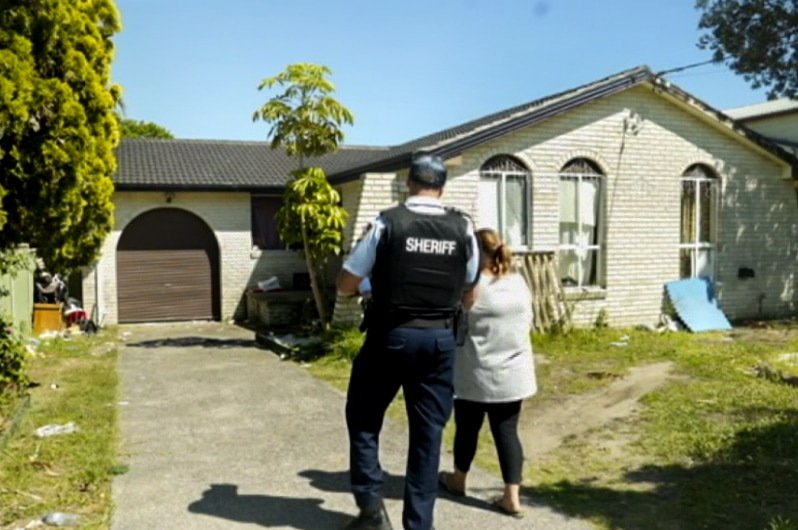 Man in blue and black uniform with Sheriff written on back walks next to a woman down a driveway toward a cream brick house