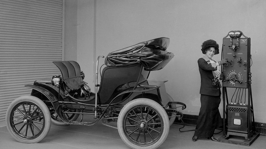 A woman with a carriage-like automobile connects a cord to a charging station and poses for the camera