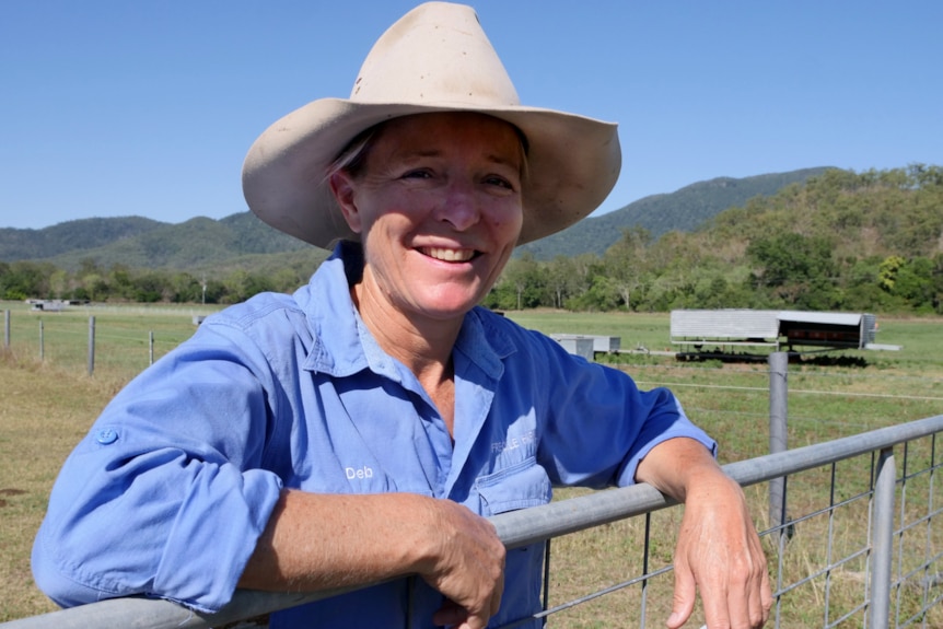 A lady wearing a broad rimmed hat is leaning on a gate and smiling at the camera.