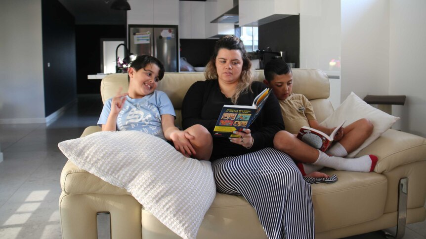 A woman sits between her two young sons on a yellow couch, reading books.