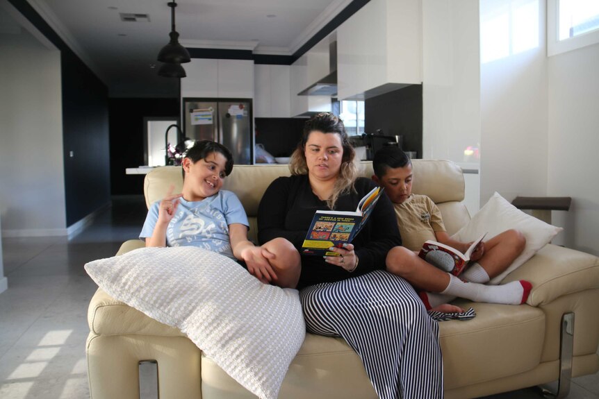 A woman sits between her two young sons on a yellow couch, reading books.