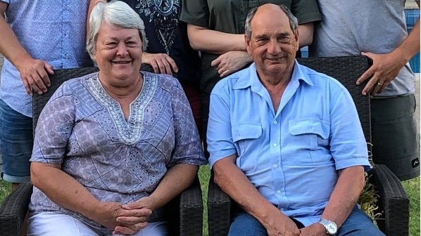 A lady and man sit in chairs side by side on a lawn with people gathered behind them. They are smiling.