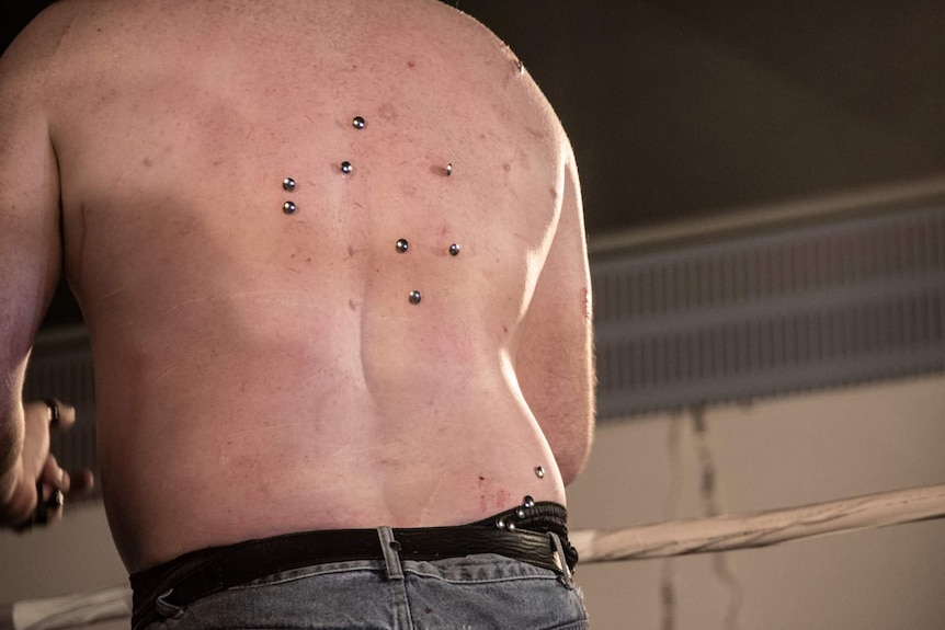 A close-up of a man's back with several silver thumb tacks stuck into the scratched and reddened skin.