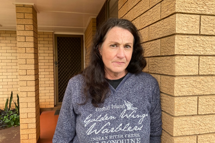 A woman with long brown hair and a navy hoodie stands in front of her brick home