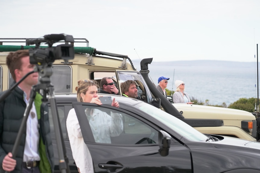 People standing by their cars looking to the right of the camera with water in the background