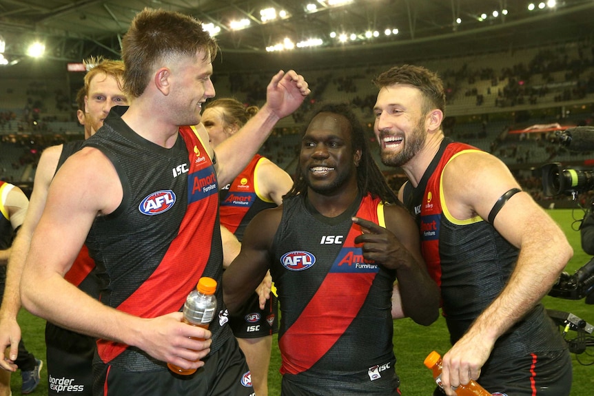 Three male AFL players smile and laugh alongside each other as they celebrate winning a match.