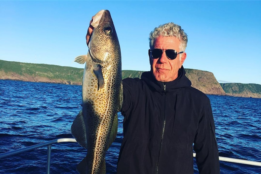 Anthony Bourdain holds up a long fish on a boat.