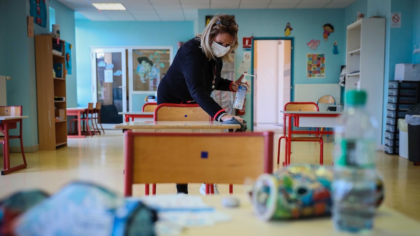 A woman wearing a face masks cleans a desk in a primary school classroom
