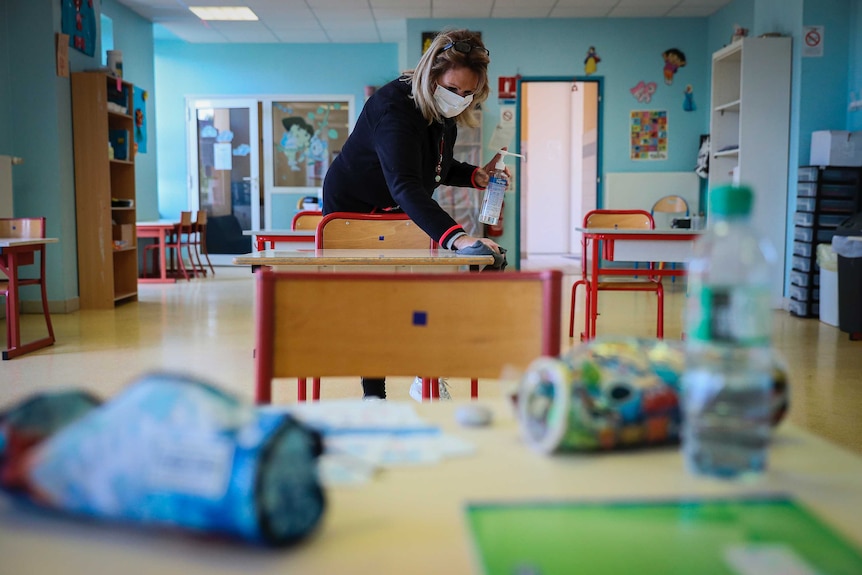 A woman wearing a face masks cleans a desk in a primary school classroom