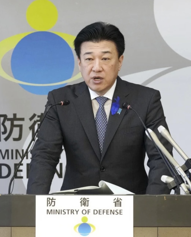 A Japanese man wearing a black suit speaks into a microphone